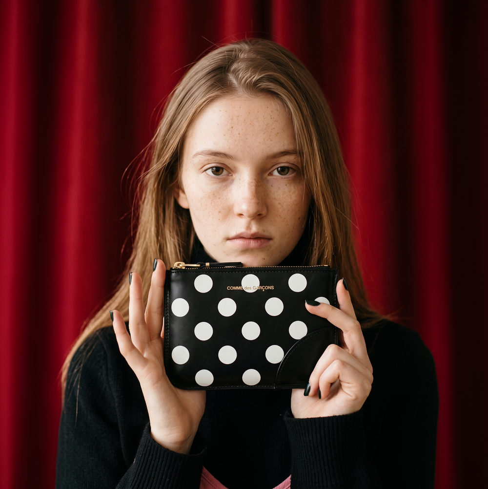 Model holding a COMME des GARÇONS black polka-dot leather zip wallet against red curtains.